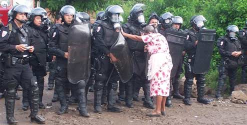 an elderly El Salvadorian women stands up to a line of riot police in armour